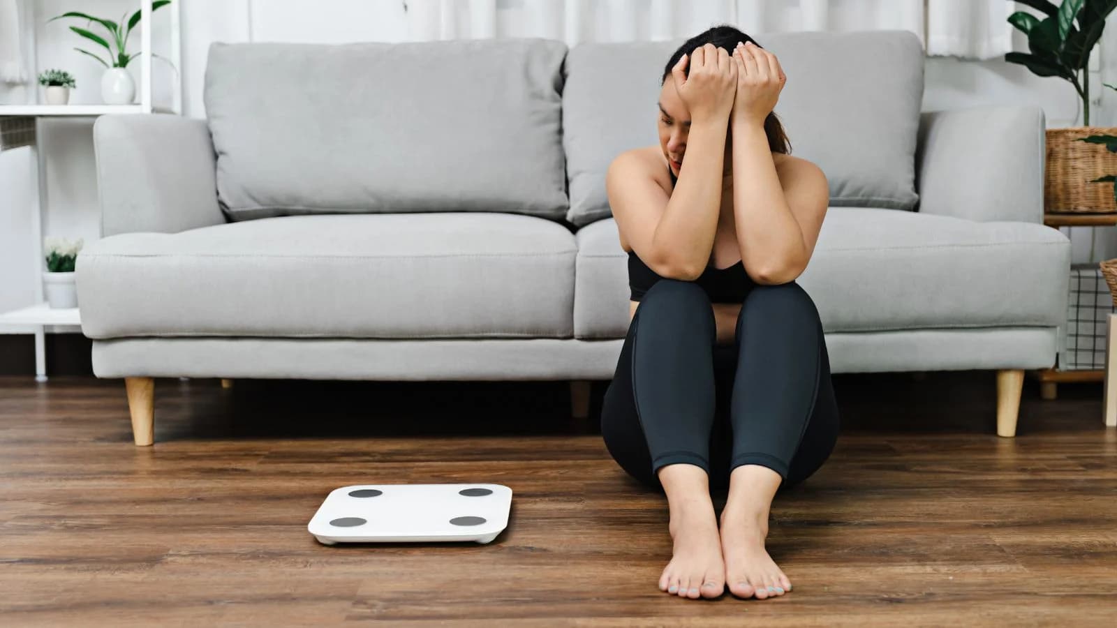 A frustrated woman sitting on the floor beside a bathroom scale after OTC weight loss products failed to work